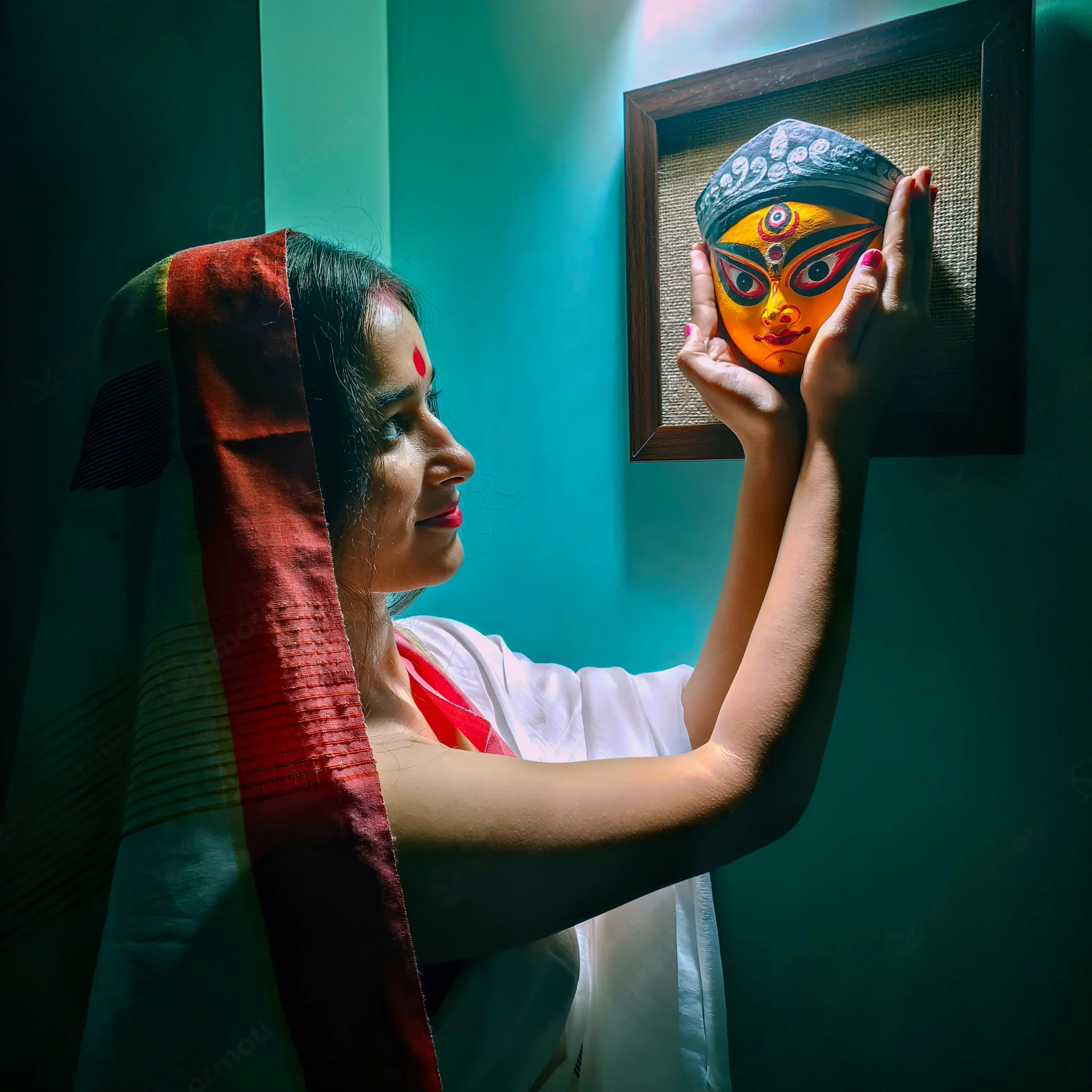 A bengali women looking at a vibrant handmade Terracotta Durga Wall Hanging on a jute background, showcasing intricate details of Maa Durga's face, a prime example of Bengal crafts.